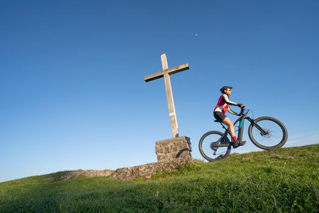 pretty senior woman riding her electric mountain bike in warm dawn sunlight below the summit cross of Salmas hight above Oberstaufen, Allgau Alps, Bavaria Germanyの写真素材