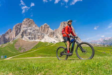pretty beautiful senior woman riding her electric mountain bike below famous Sassolungo summit in Sella Dolomites of Selva Wolkenstein, Val Gardena, South Tirol and Trentino, Italyの写真素材