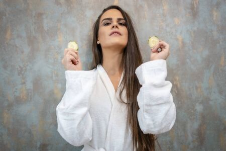 A young brunette in a white coat in a beauty salon with cucumbers in her handの写真素材