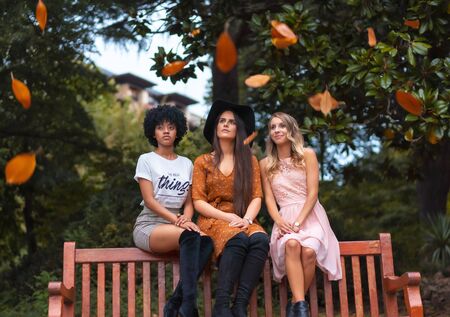 Street style session. Three friends sitting in the park in autumn and falling red leaves, a blonde, a brunette and a Latin girl with afro hairの写真素材
