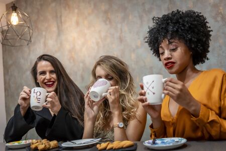Session in a cafeteria. Three young girlfriends drinking coffee in a cafeteria. Blonde girl, brunette girl and girl of Caribbean American origin with afro hairの写真素材
