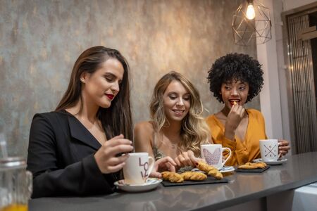 Session in a cafeteria. Three very happy friends having coffee and some pastries in a cafeteria. Blonde girl, brunette girl and girl of Caribbean American origin with afro hairの写真素材