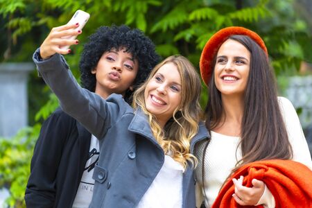 Street style session. Three smiling friends making a selfie in the city, a blonde, a brunette and a Latin girl with afro hairの写真素材