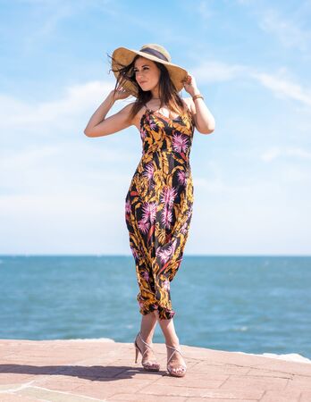 Street style, posing of a slim young brunette with a beautiful floral dress and a hat with the sea in the backgroundの写真素材