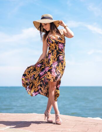 Street style, posed by a pretty young brunette in a beautiful floral dress moved by the wind and a large straw hat. With the sea in the backgroundの写真素材