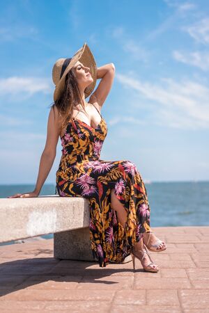 Street style, a young brunette in a beautiful floral dress sitting on the coast in summer enjoying the sunの写真素材