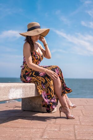 Lifestyle, a pretty young brunette sitting in a floral dress and a straw hat by the seaの写真素材
