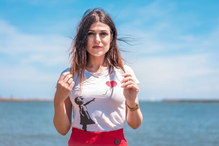 Street style, portrait of young serious brunette in white t-shirt and red skirt on the coast with the sea in the backgroundの写真素材