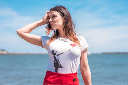 Street style, portrait of young brunette in white shirt and red skirt on the coast with the sea in the background looking to the leftの写真素材