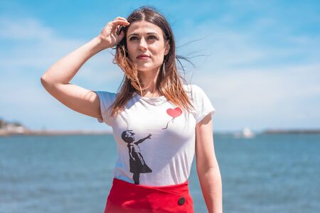 Street style, portrait of young brunette in white shirt on the coast with the sea in the background looking to the leftの写真素材