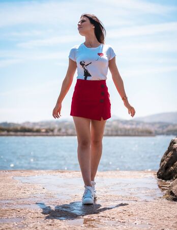 Street style, portrait of a pretty brunette in a white t-shirt, a red short skirt and white sneakers with the sea behindの写真素材