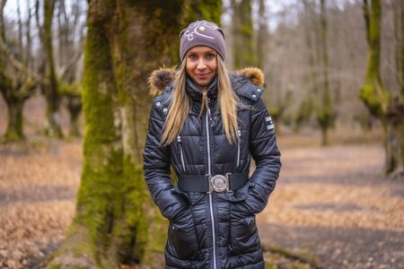Lifestyle, portrait of smiling blonde Caucasian woman between trees in a forest with black jacket in winterの写真素材