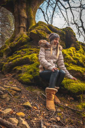 A young blonde woman sitting on the roots of a tree with moss