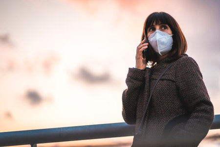 A young brunette talking on the phone with a mask on a sunset. First walks of the uncontrolled...