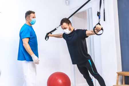Physiotherapist with mask and a patient doing stretches upside down with rubber bands. Physiotherapy with protective measures for the Coronavirus pandemic, COVID-19. Osteopathy, sports chiromassageの写真素材