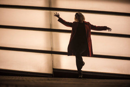 Silhouette of a young blonde woman with open arms leaning against a bright wall in the city, street...