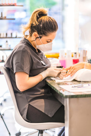 A young Latina worker at a manicure and pedicure salon with security measures and face masks. The...