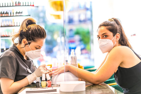 Latin girl with face mask painting colored nails to the Caucasian brunette client. Reopening after...