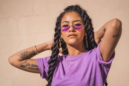 Urban session. Young dark-skinned woman with long braids wearing purple glasses on a plain cream-colored background, with serious lookの写真素材
