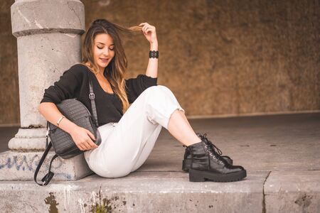 Lifestyle in the city with a blonde girl in white pants and black wool sweater near the beach. Sitting on the ground next to the seafrontの写真素材