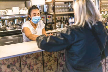 Photo session with a waitress with a face mask in a bar. New normality, security measures after the coronavirus pandemic, social distance, covid-19. Delivering a coffee to a clientの写真素材