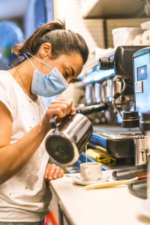 Photo session with a waitress with a face mask in a bar. New normality, security measures after the coronavirus pandemic, social distance, covid-19. Throwing milk into a cup of coffeeの写真素材