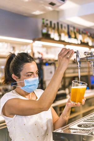 Photo session with a waitress with a face mask in a bar. New normality, security measures after the coronavirus pandemic, social distance, covid-19. Putting a cold beerの写真素材