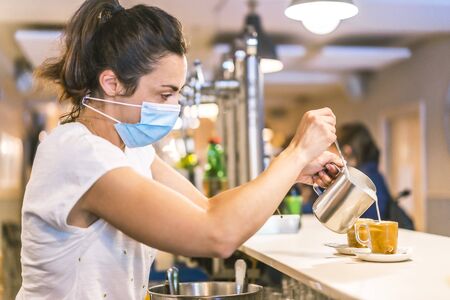 Photo session with a waitress with a face mask in a bar. New normality, security measures after the coronavirus pandemic, social distance, covid-19. Putting milk in a cup of coffeeの写真素材