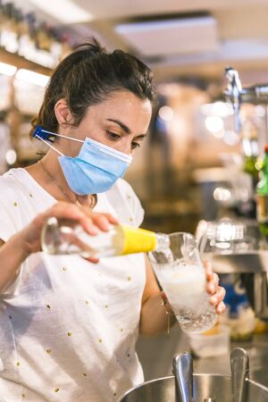Photo session with a waitress with a face mask in a bar. New normality, security measures after the coronavirus pandemic, social distance, covid-19. Putting a gin and tonic in a glassの写真素材