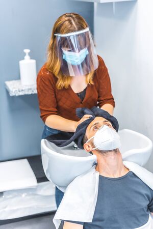 Reopening of hairdressing salons after the Coronavirus pandemic. Hairdresser with face mask and protective screen, covid-19. Social distance, new normality. Pulling client's hair out with mascaraの写真素材