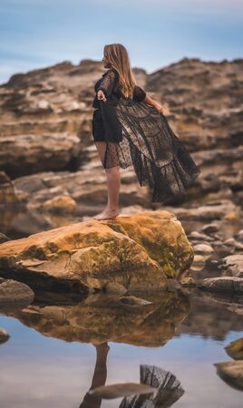 Summer lifestyle with a young brunette Caucasian woman in a long black transparent dress on some rocks near the sea on a summer afternoon. Raising your arms on top of a rockの写真素材