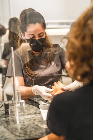 Manicure and pedicure salon, coronavirus, covid-19, social distance. The reopening due to the pandemic, security measures. A worker and a client with masks in a treatment with a protective screenの写真素材