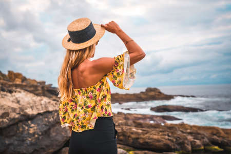 Caucasian blonde girl in a floral shirt, black shorts and straw hat in a natural landscape by the sea and rocks at sunset, lifestyle. Posing standing by the seaの写真素材