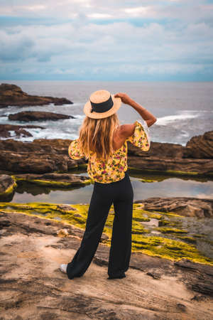 Caucasian blonde girl in a floral shirt, black shorts and straw hat in a natural landscape by the sea and rocks at sunset, lifestyle. Enjoying the summer afternoonの写真素材