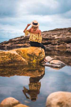 Caucasian blonde girl in a floral shirt, black shorts and straw hat in a natural landscape by the sea and rocks at sunset, lifestyle. Sitting on a rock with your back to you and lookingの写真素材