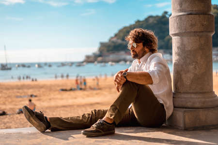 Summer lifestyle with a young dark-haired Caucasian man with long beard, sunglasses, shirtless in a white shirt open on a beach. San Sebastian, Basque Country. Sitting looking at the seaの写真素材