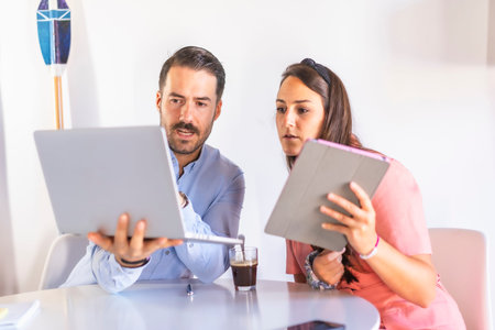 A couple teleworking at home, boy in blue shirt and girl in pink suit. With a laptop and tablet from...