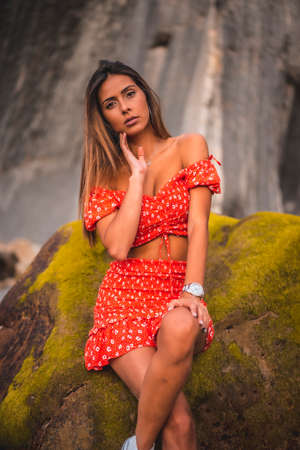 A young brunette Caucasian woman in a red dress on the beach of Itzurrun in the town of Zumaia, Gipuzkoa. Basque Country. Lifestyle session, portrait sitting on a green sea rockの写真素材