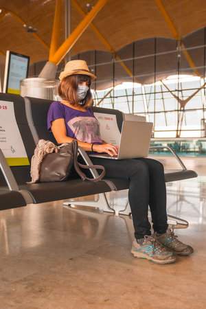 Air travel in the coronavirus pandemic, safe travel, social distance, new normal. A young woman with a face mask working with a computer waiting to take off, almost empty airportsの写真素材