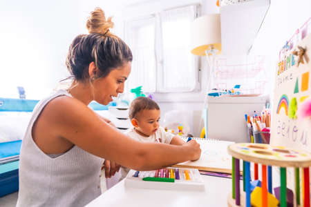 Young Caucasian mother playing with her in the room with toys. Baby less than a year learning the first lessons of her mother. Painting with color paintsの写真素材