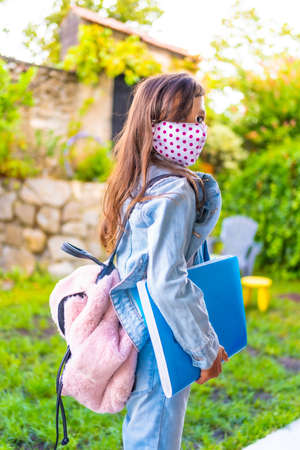 Caucasian girl with face mask ready to go back to school. New normality, social distance, coronavirus pandemic, covid-19. Jacket, backpack, mask with pink dots and a blue block in handの写真素材