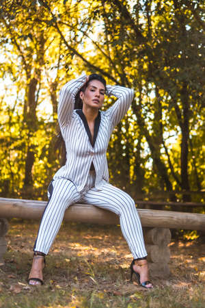 Summer lifestyle, young Latin brunette girl in a very elegant white suit with black stripes. Fashion posed in a beautiful park sitting, fashion editorialの写真素材
