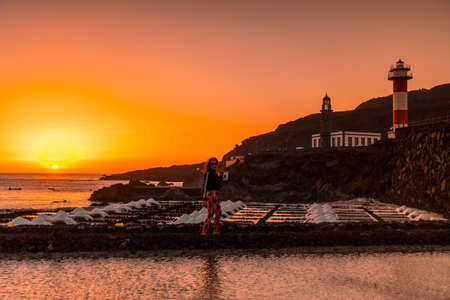 A young woman walking in the orange sunset in the salina and in the background the Fuencaliente Lighthouse on the route of the volcanoes south of the island of La Palma, Canary Islands, Spainの写真素材