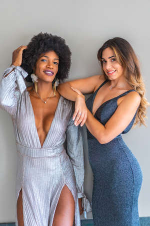 Two multi-ethnic friends in ball gowns ready to go out at night to the exclusive party. Gray and blue evening dresses, blonde caucasian girl, black girl with afro hair, gray wall backgroundの写真素材