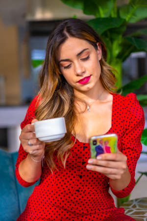 Caucasian girl in a red dress having a coffee and writing a message on the phone. Sitting on a blue sofa, young enterprising woman in a hotelの写真素材