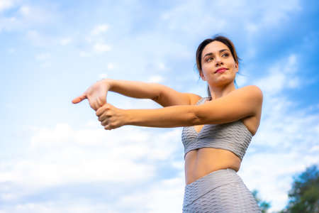 Fitness session with a young Caucasian blonde, very cheerful doing stretching, with the sky in the background, exercising in the field, gray sport suit, fit girl, healthy lifeの写真素材