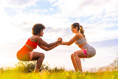 Caucasian blonde girl and dark-skinned girl with afro hair doing squat exercises in a park with the city in the background. Healthy life, fitness, fitness girls, gray and red sport outfitsの写真素材
