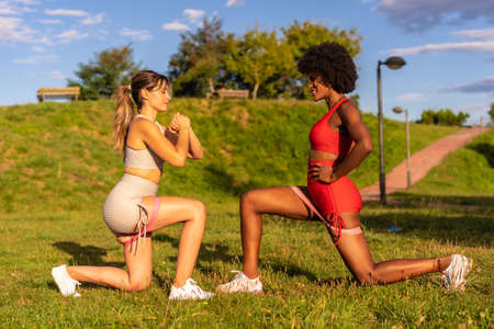 Caucasian blond girl and dark-skinned girl with afro hair doing stretching exercises in a park. Healthy life, fitness, fitness girls, gray and red sport outfitsの写真素材