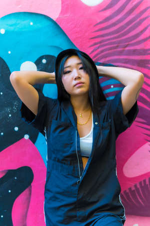 Young Asian rapper girl in a black cap and black jumpsuit crouched on the ground in the city, photo trap pose.の写真素材