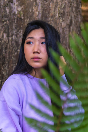 Portrait of a young Asian brunette in a park next to a tree with a pink sweater.